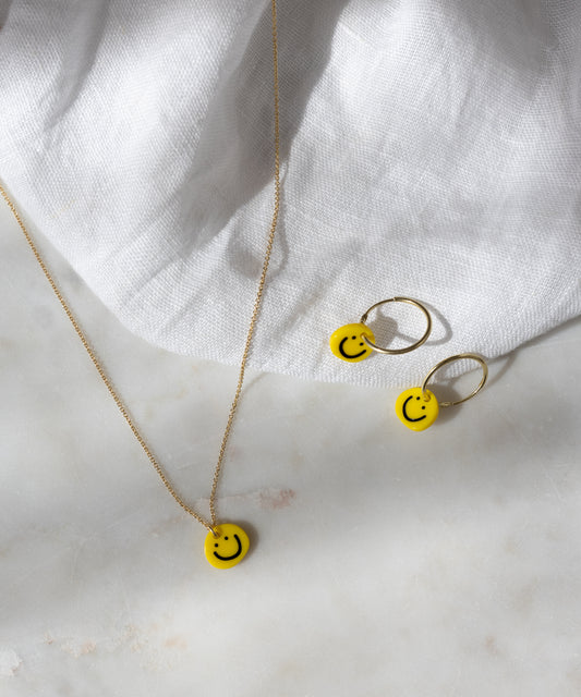 Smilie Yellow Earrings by WALD Berlin, featuring 18k gold-plated hoops with yellow smiley face charms, displayed on a white fabric and marble surface.