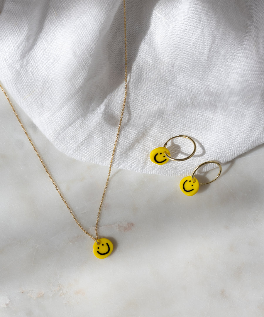 Smilie Yellow Earrings by WALD Berlin, featuring 18k gold-plated hoops with yellow smiley face charms, displayed on a white fabric and marble surface.