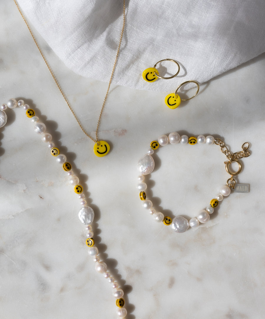 Eine Perlenkette und ein Armband, verziert mit gelben Smiley-Perlen, eine goldene Kette sowie die „Smilie Yellow“-Ohrringe von WALD Berlin – vergoldete Creolen mit passenden, in Deutschland gefertigten Smiley-Anhängern – präsentiert auf einer weißen Marmorfläche.