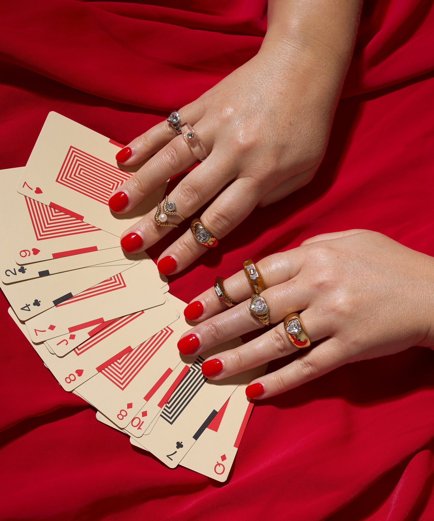 Two hands with red nail polish and several WALD Berlin Be My Lover Mini Silver Rings hold a fan of playing cards against a red fabric backdrop.
