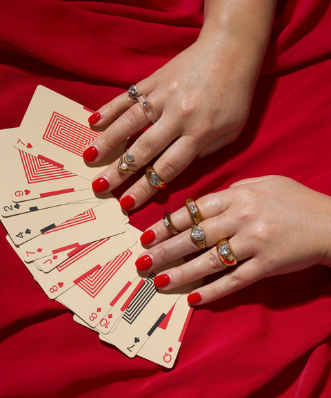 Two hands with red nail polish wear WALD Berlin's NYC If I Rule The World Ring, displaying a fanned-out deck of playing cards on a red fabric background.