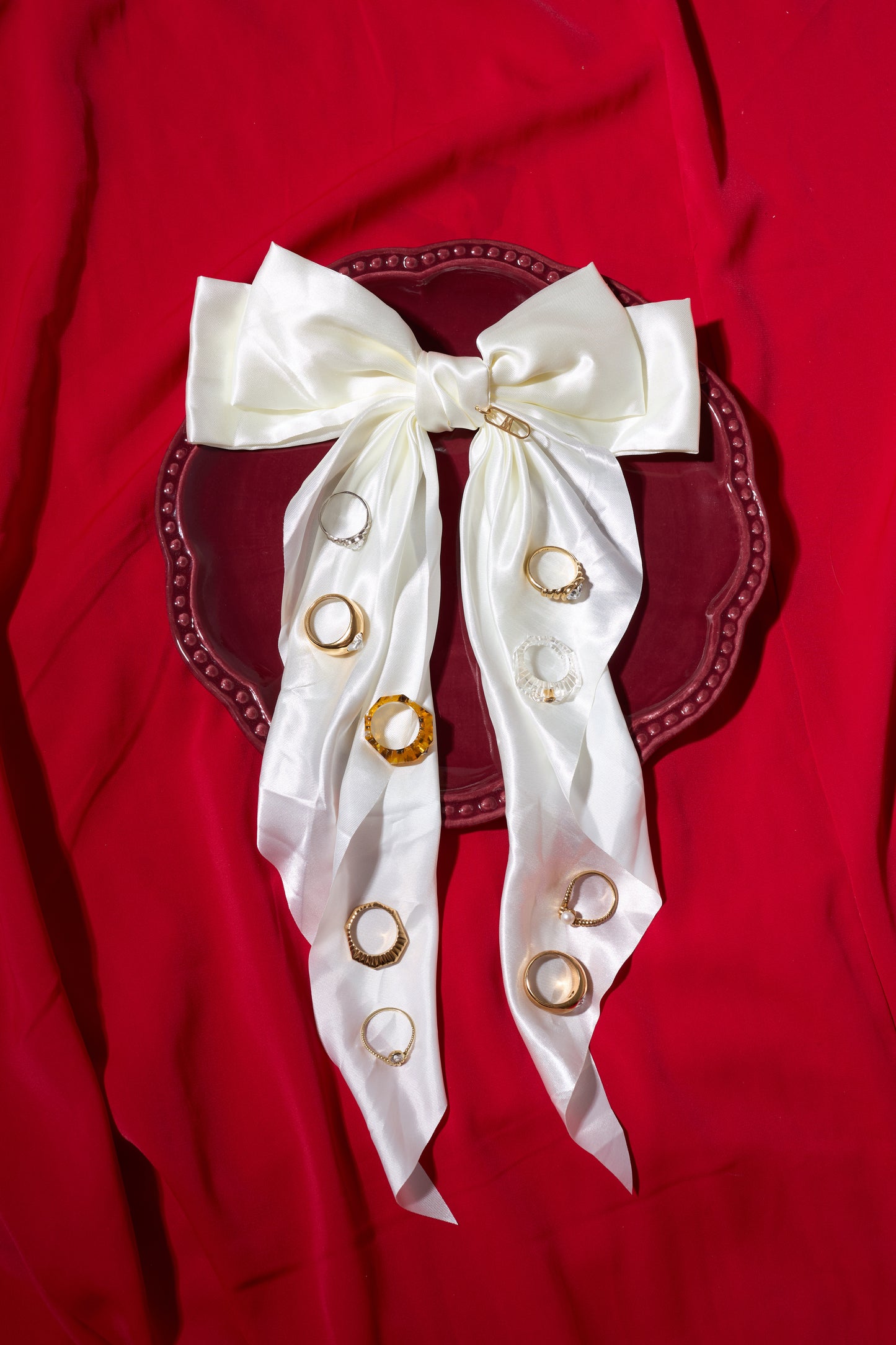 The WALD Berlin NYC If I Rule The World Ring, handmade in Germany, is shown on a maroon plate with a white satin bow and gold-plated accents, set against a red fabric background.