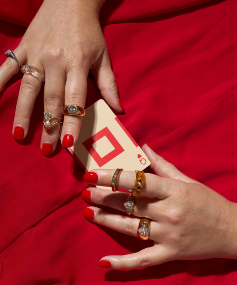Two hands with red nails wear multiple WALD Berlin NYC If I Rule The World Rings, holding a playing card with a red geometric design against a red fabric background.