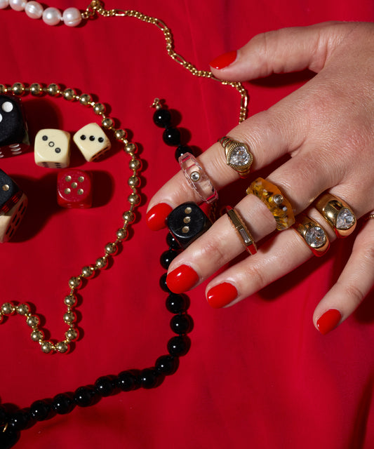 A hand with red nail polish wears multiple rings, including the Pop Swarovski Ring by WALD Berlin, and rests on a red surface surrounded by necklaces and various dice.