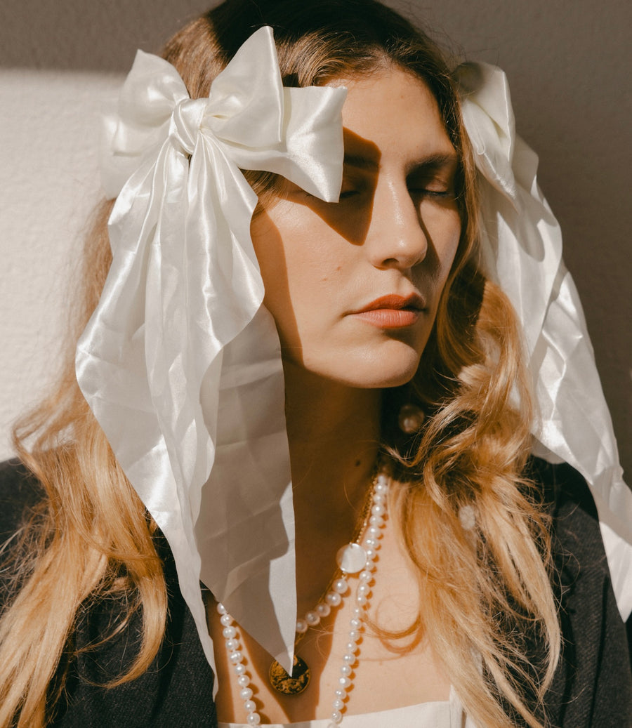 A woman with long hair, eyes closed, is adorned in festive styling featuring WALD Berlin's Happy Holidays Cream Hair Bow and pearl necklaces against a light background.
