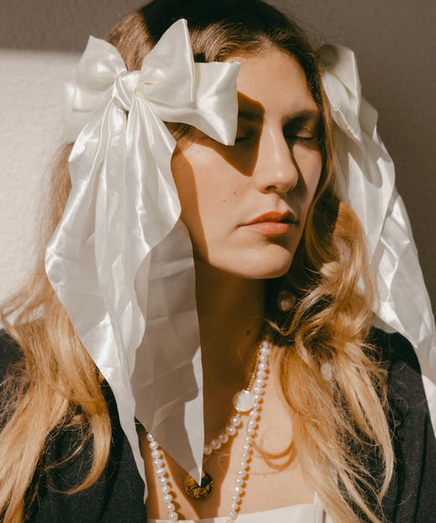 Under soft lighting, a woman with long hair peacefully closes her eyes, capturing the essence of festive styling as she wears a large Happy Holidays Black Hair Bow from WALD Berlin and a pearl necklace.