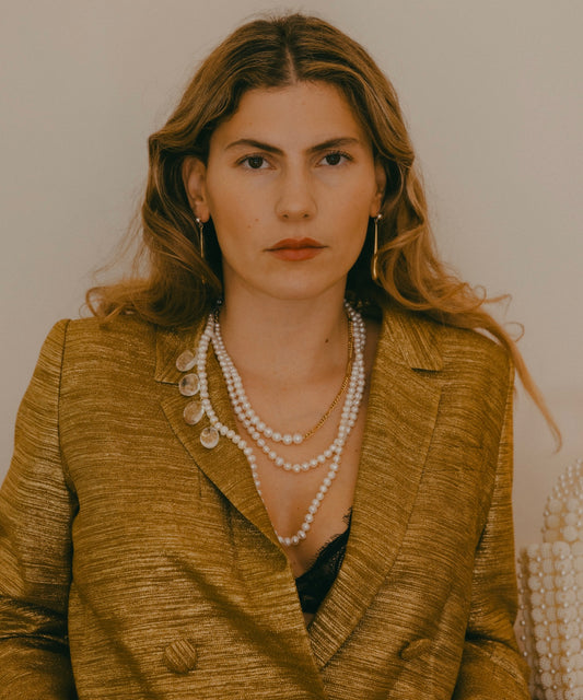 A woman with wavy hair is wearing a gold jacket, WALD Berlin's Happy Holidays Long Pearl Drop Necklace, and hoop earrings while looking directly at the camera against a plain background.