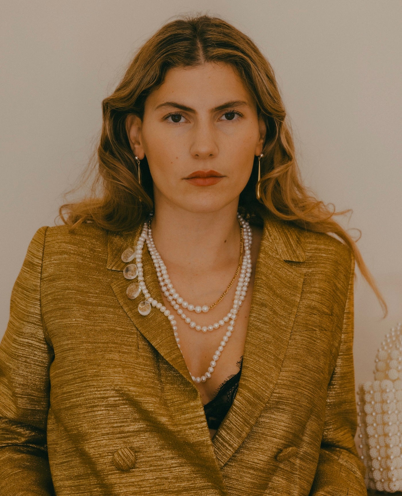 A woman with wavy hair is wearing a gold jacket, WALD Berlin's Happy Holidays Long Pearl Drop Necklace, and hoop earrings while looking directly at the camera against a plain background.