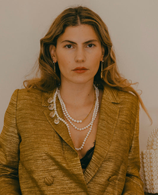 A woman with wavy hair is wearing a gold jacket, WALD Berlin's Happy Holidays Long Pearl Drop Necklace, and hoop earrings while looking directly at the camera against a plain background.