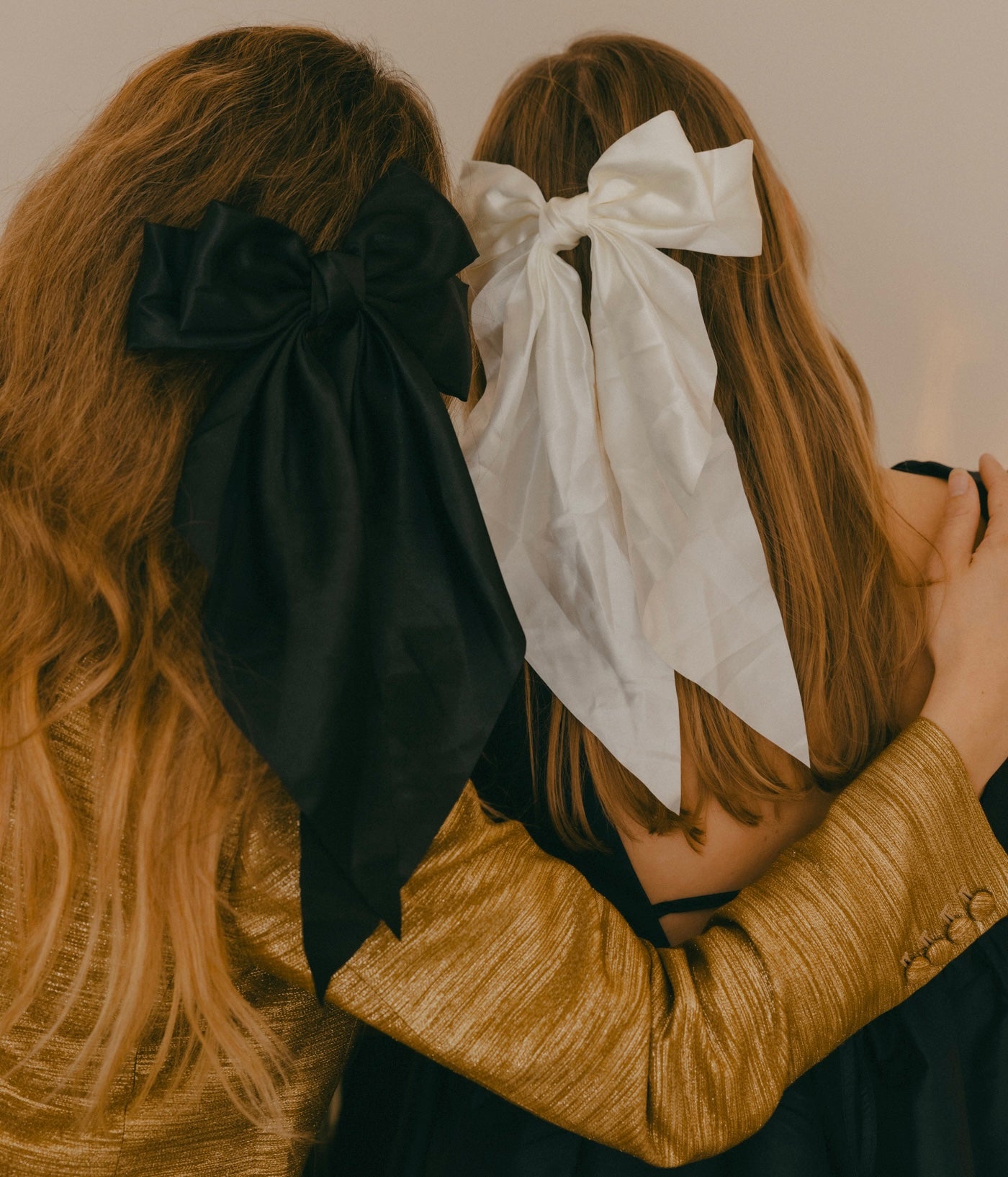 Two individuals with long hair, each highlighting a festive hair accessory—one featuring WALD Berlin's "Happy Holidays Black Hair Bow" and the other in white—bring a festive flair to their holiday hairstyles. The person on the left catches the eye with a shimmering gold top, ideal for the season's parties and gatherings.