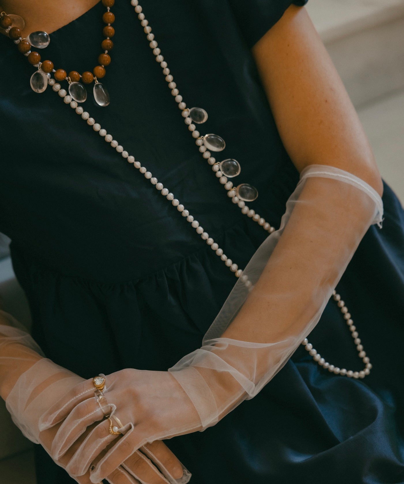 A person in a navy dress with sheer gloves sits with arms crossed, adorned with layered necklaces, including the "Happy Holidays Long Pearl Drop Necklace" by WALD Berlin, featuring berg crystal pendants and sweetwater pearls, complemented by multiple rings.