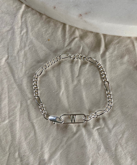 The Naomi Bracelet Silver by WALD Berlin features a decorative safety pin with the letter "W," crafted from recycled gold and silver, and is shown on beige textured fabric beside a white stone.