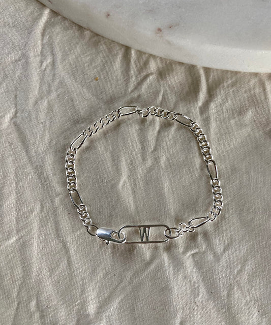 The Naomi Bracelet Silver by WALD Berlin features a decorative safety pin with the letter "W," crafted from recycled gold and silver, and is shown on beige textured fabric beside a white stone.