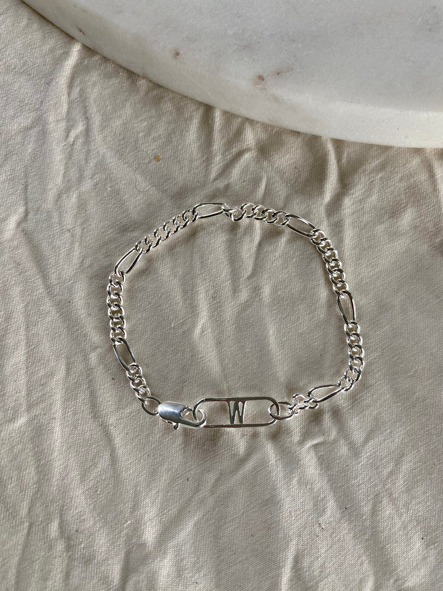 The Naomi Bracelet Silver by WALD Berlin features a decorative safety pin with the letter "W," crafted from recycled gold and silver, and is shown on beige textured fabric beside a white stone.