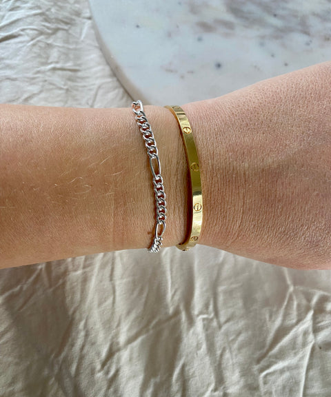 A wrist adorned with WALD Berlin's Naomi Bracelet Silver, a thin recycled silver chain, alongside a gold engraved bangle, rests on light fabric—an elegant touch of German jewellery.