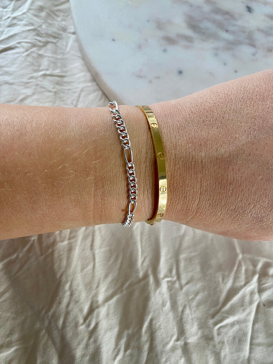A wrist adorned with WALD Berlin's Naomi Bracelet Silver, a thin recycled silver chain, alongside a gold engraved bangle, rests on light fabric—an elegant touch of German jewellery.