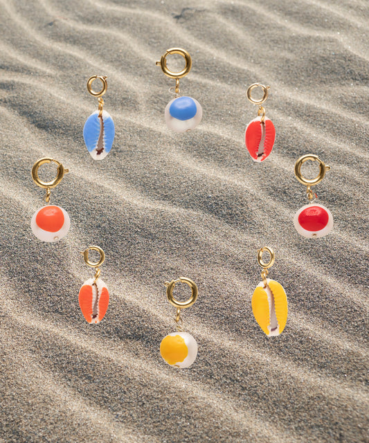 Nine gold-plated brass earrings from WALD Berlin's SAMPLE SALE Beach Babe Charms, featuring vibrant cowrie shells and circular pendants in blue, red, orange, and yellow, are artfully arranged on rippled sand.