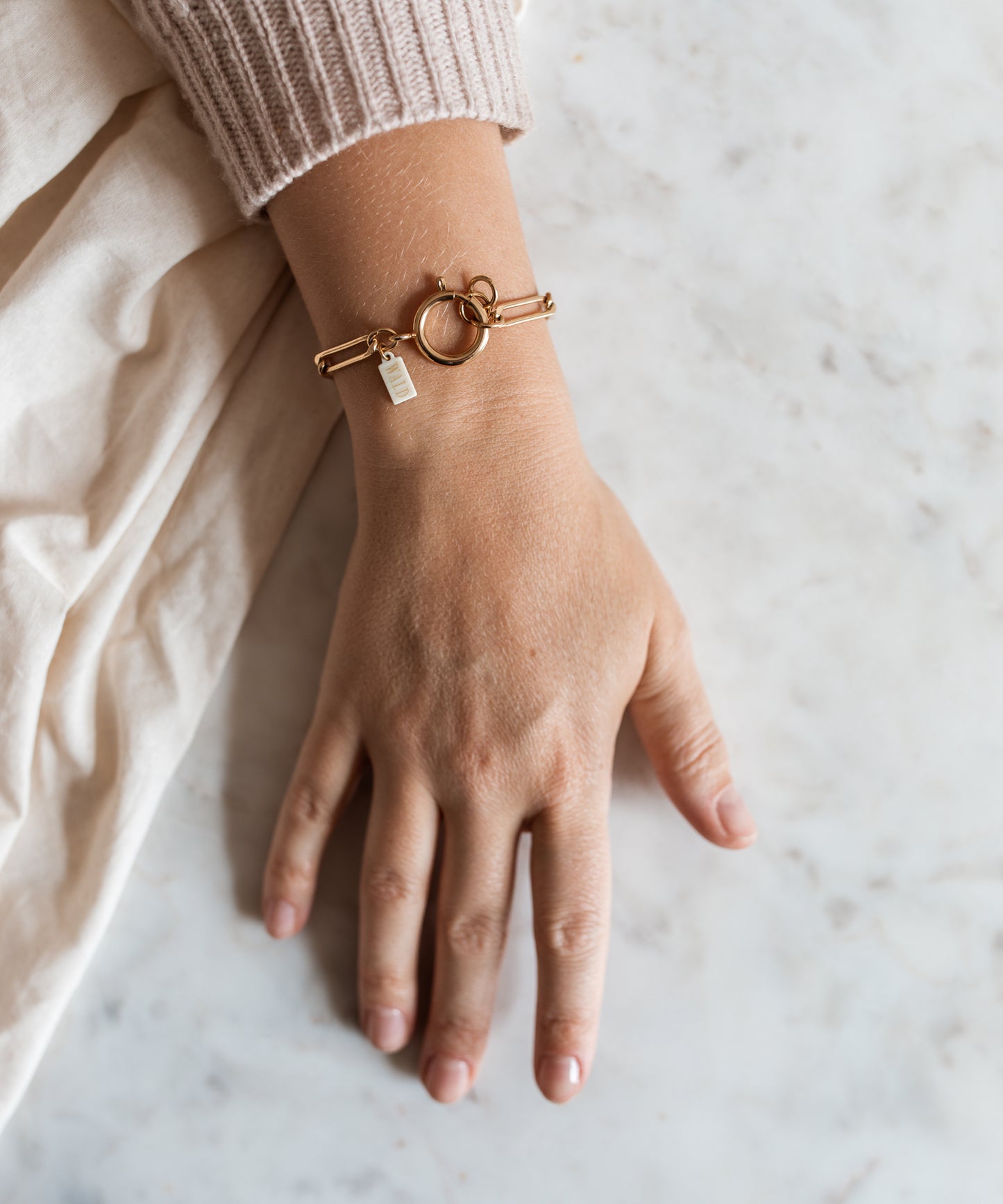 A hand on a marble surface wears the SAMPLE SALE Ashley Gold Bracelet by WALD Berlin, featuring a circular clasp and small rectangular charm, partially tucked beneath a beige sweater sleeve.