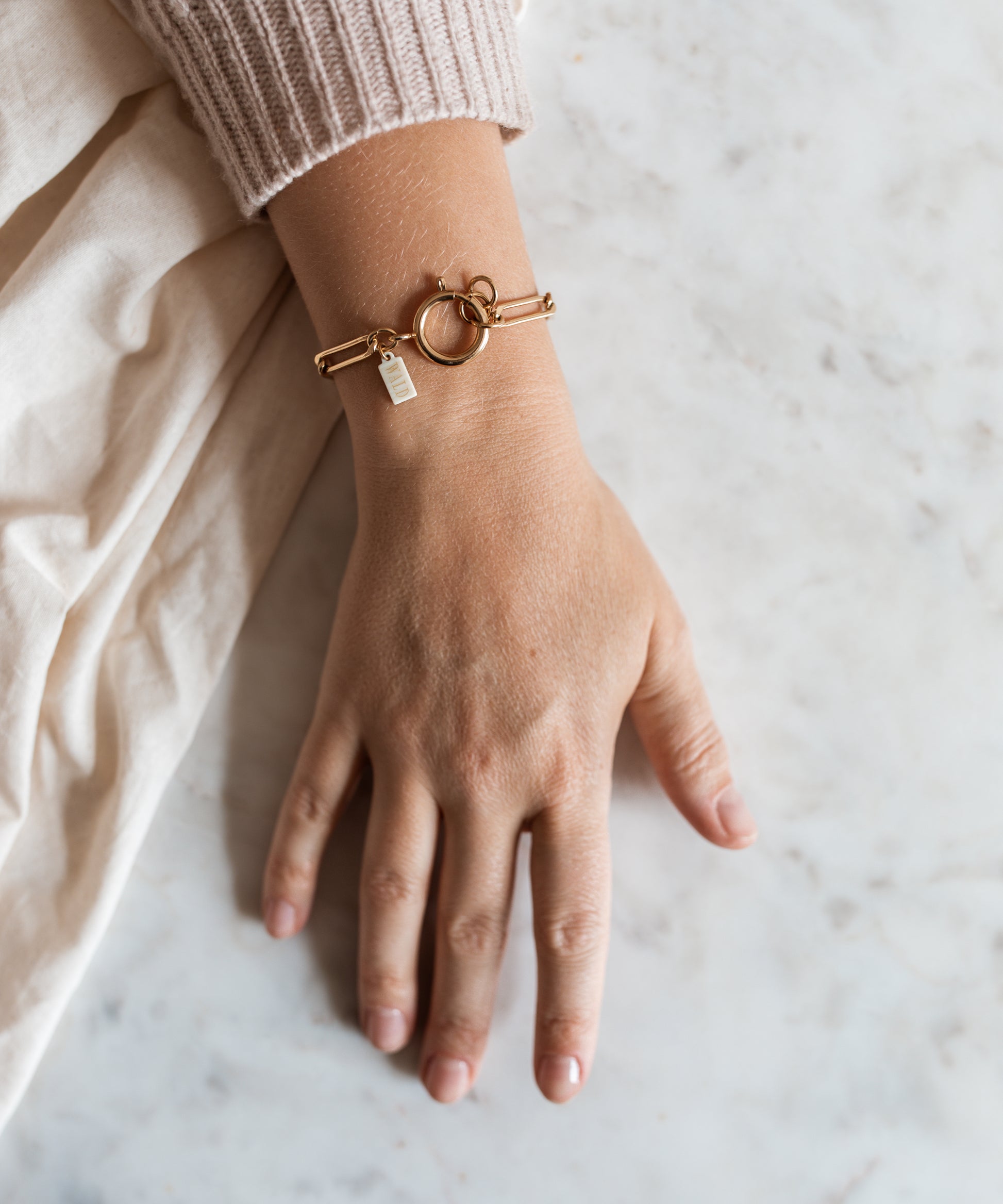 A hand on a marble surface wears the SAMPLE SALE Ashley Gold Bracelet by WALD Berlin, featuring a circular clasp and small rectangular charm, partially tucked beneath a beige sweater sleeve.