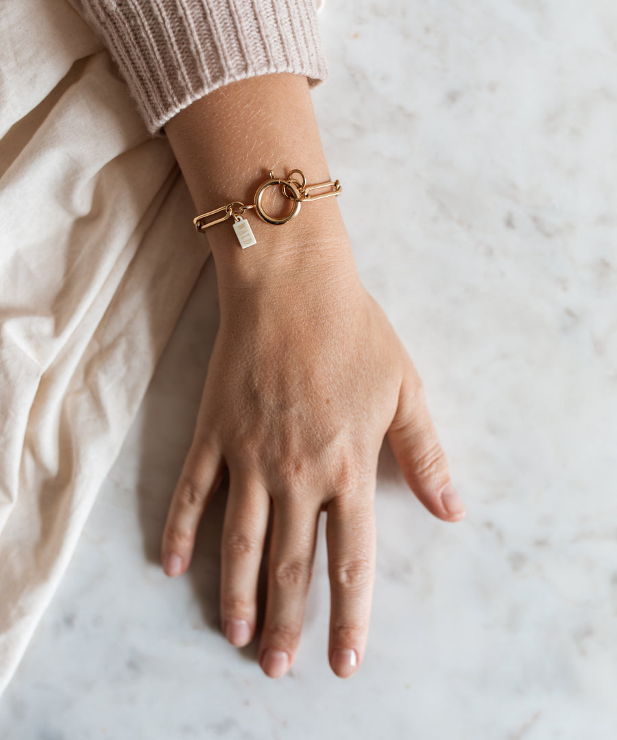 A hand on a marble surface wears the SAMPLE SALE Ashley Gold Bracelet by WALD Berlin, featuring a circular clasp and small rectangular charm, partially tucked beneath a beige sweater sleeve.
