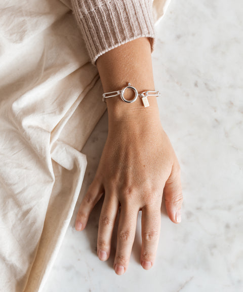 A hand wearing the WALD Berlin SAMPLE SALE Ashley Bracelet Silver, featuring a circular clasp and small rectangular charm, rests on a marble surface beside beige fabric.