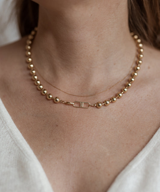 A close-up of a person wearing the Harry Necklace Gold by WALD Berlin, a gold-plated brass necklace featuring a small rectangular pendant with the letter "W," elegantly draped on a thin chain. The German jewelry piece beautifully complements their light-colored top.