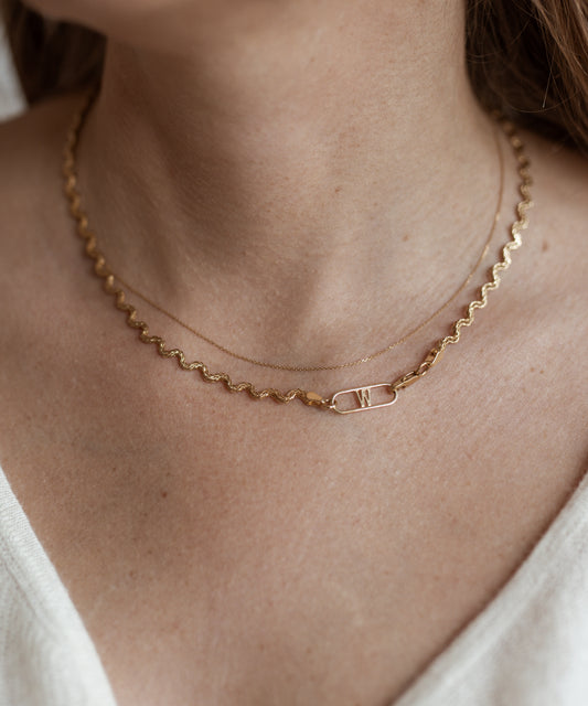 Close-up of a person wearing two necklaces, including the Irina Necklace Gold from WALD Berlin: a wavy gold chain and a delicate recycled gold chain with a rectangular pendant featuring the "W" initial.