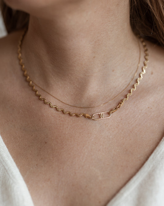 Close-up of a person wearing two necklaces, including the Irina Necklace Gold from WALD Berlin: a wavy gold chain and a delicate recycled gold chain with a rectangular pendant featuring the "W" initial.