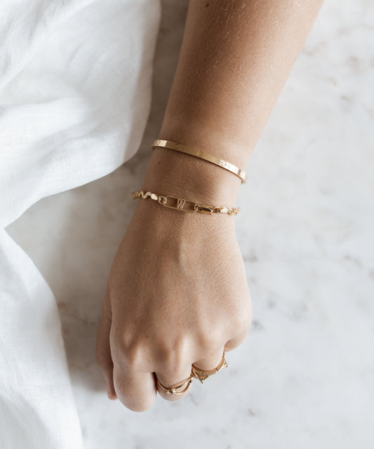 Adorning a hand that rests elegantly on a marble surface beside white fabric, two Irina Gold Bracelets and three gold rings exemplify the exquisite craftsmanship of WALD Berlin, all expertly made in Germany.