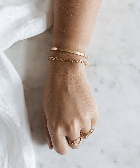 A hand adorned with an Irina Gold Bracelet by WALD Berlin and matching rings rests elegantly on a light marble surface, showcasing timeless craftsmanship.