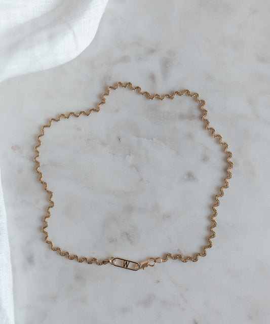 The Irina Necklace Gold by WALD Berlin, made from recycled gold with a wavy chain design and a clasp adorned with an engraved letter 'W', is gracefully showcased on a white marble surface.