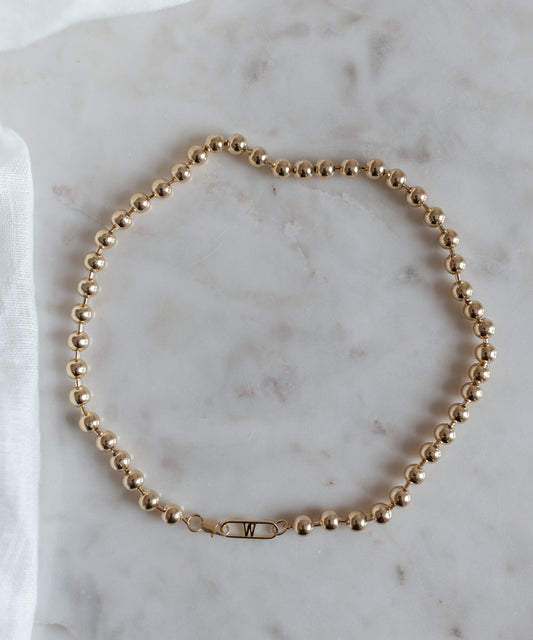 The WALD Berlin Harry Necklace Gold, a gold-plated brass beaded necklace featuring a clasp with the letter "W," is elegantly arranged on a marble surface.