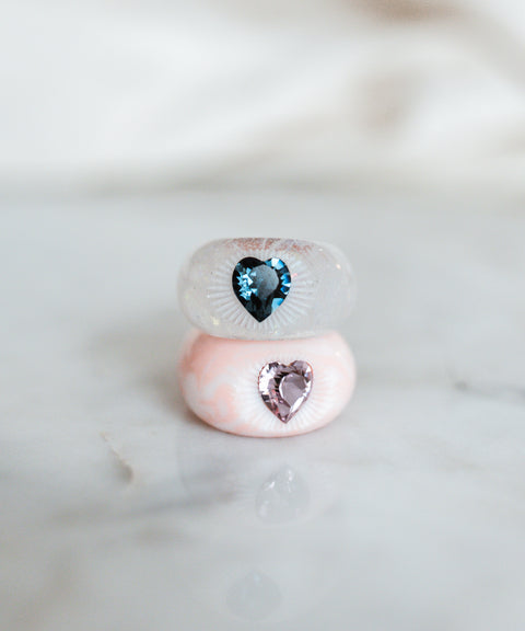 A pair of WALD x Zeena Rose Heart Rings, one in pink and the other in blue, adorned with Swarovski stones, placed delicately on a marble table.