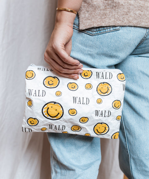 A woman holding a yellow Smilie Dude Pouch makeup bag by WALD Berlin, a trendy jewelry brand from Germany.
