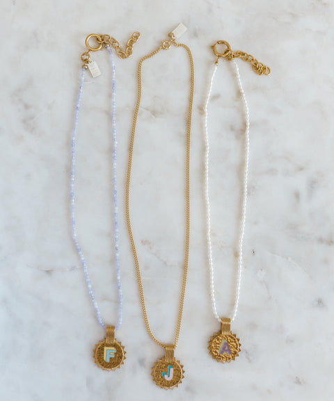 Three WALD Berlin Sample saleSay My Name Gold Necklaces feature gold-plated round initial pendants (F, J, A) with adjustable chains in light blue, gold, and white, elegantly displayed on a marble surface.