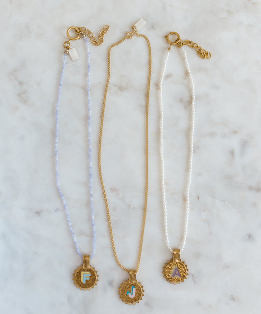 Three WALD Berlin Sample saleSay My Name Gold Necklaces feature gold-plated round initial pendants (F, J, A) with adjustable chains in light blue, gold, and white, elegantly displayed on a marble surface.