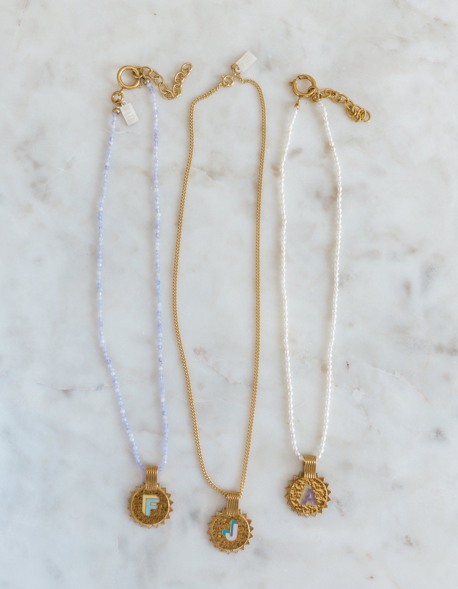 Three WALD Berlin Sample saleSay My Name Gold Necklaces feature gold-plated round initial pendants (F, J, A) with adjustable chains in light blue, gold, and white, elegantly displayed on a marble surface.