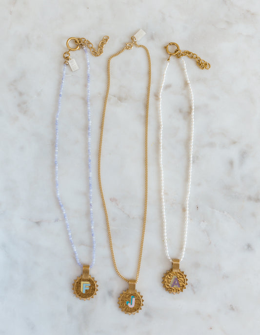 Three WALD Berlin Sample saleSay My Name Gold Necklaces feature gold-plated round initial pendants (F, J, A) with adjustable chains in light blue, gold, and white, elegantly displayed on a marble surface.