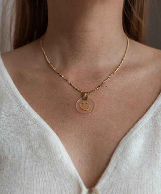 A person wearing the sophisticated Smilie Dude Medaillon Gold Necklace by WALD Berlin, featuring an 18k gold-plated finish and a transparent pendant with a cheerful yellow smiling face, paired over a white V-neck top.