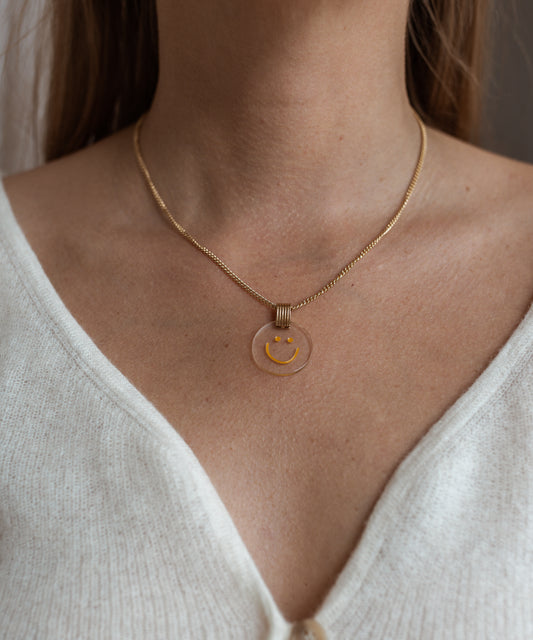 A person wearing the sophisticated Smilie Dude Medaillon Gold Necklace by WALD Berlin, featuring an 18k gold-plated finish and a transparent pendant with a cheerful yellow smiling face, paired over a white V-neck top.