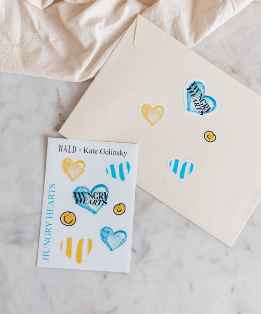 A cream envelope and a card decorated with the WALD x Kate Gelinsky Sticker Sheet showing hearts, stripes, and smiley faces—all branded under "Hungry Hearts." Inside, you'll find an elegant heart necklace that brings a sophisticated charm to this delightful package from WALD Berlin.