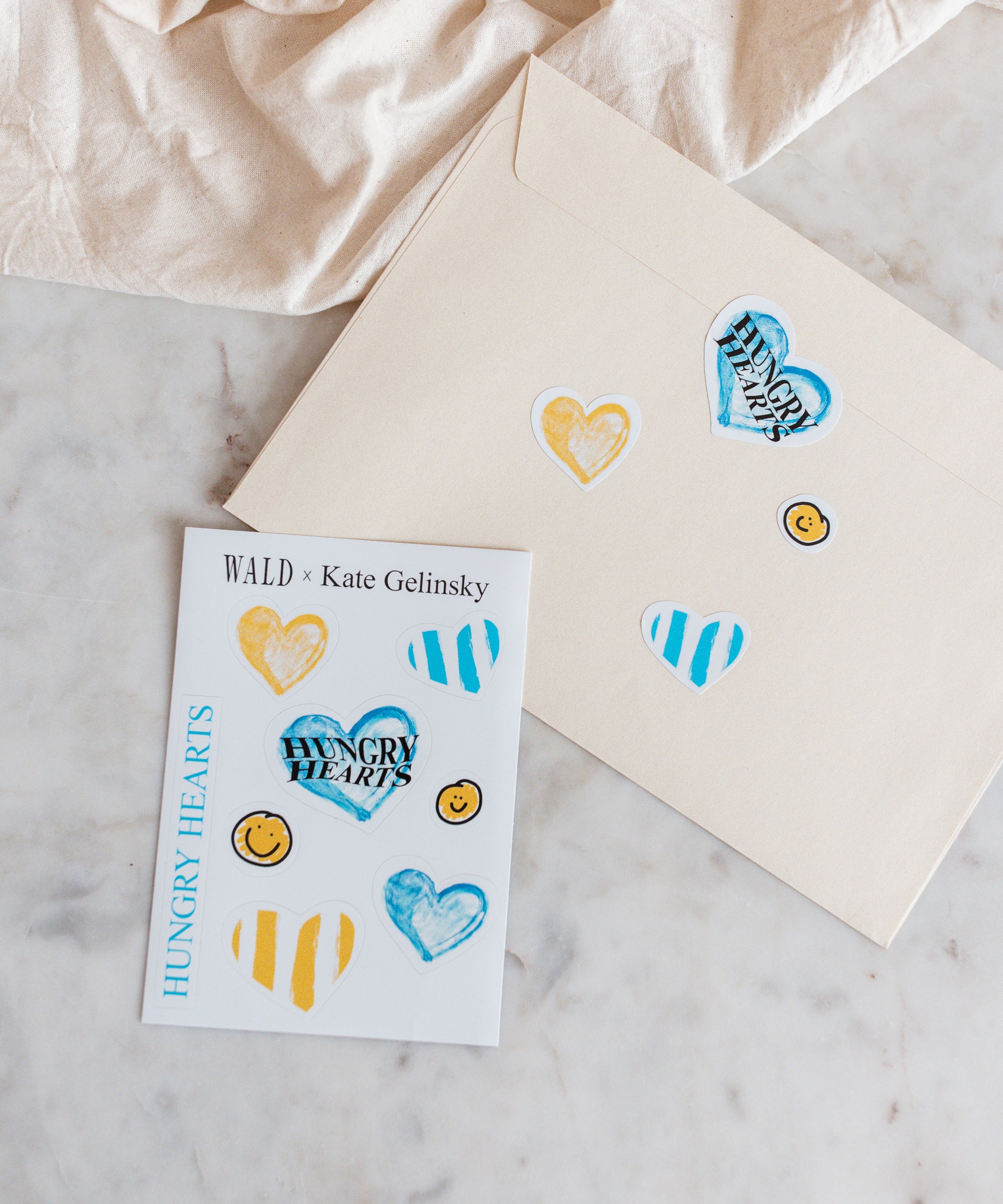 A cream envelope and a card decorated with the WALD x Kate Gelinsky Sticker Sheet showing hearts, stripes, and smiley faces—all branded under "Hungry Hearts." Inside, you'll find an elegant heart necklace that brings a sophisticated charm to this delightful package from WALD Berlin.