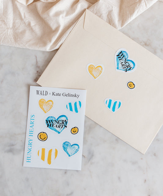 A cream envelope and a card decorated with the WALD x Kate Gelinsky Sticker Sheet showing hearts, stripes, and smiley faces—all branded under "Hungry Hearts." Inside, you'll find an elegant heart necklace that brings a sophisticated charm to this delightful package from WALD Berlin.