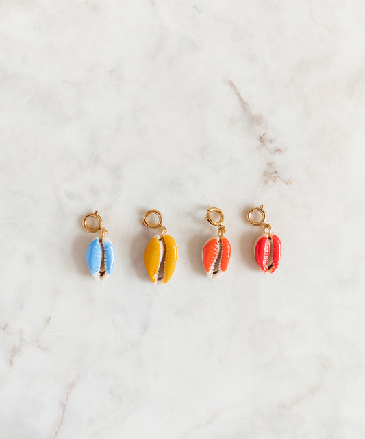 Four SAMPLE SALE Beach Babe Charms by WALD Berlin, each cowrie shell with a gold loop and painted in blue, yellow, orange, and red, are arranged in a row on a light marble surface.