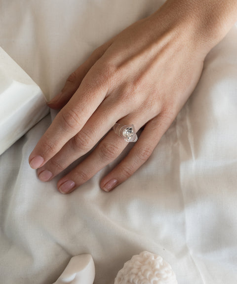 A hand with neatly trimmed nails rests on white fabric, wearing WALD Berlin's Lover Transparent Ring—a clear, heart-shaped stone delicately set on the ring finger.