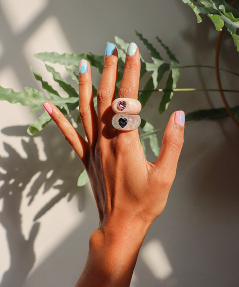 A hand with light brown skin and pastel multicolored nails displays the WALD Berlin SAMPLE Zeena x WALD Blue Heart Ring, a chunky romantic jewelry piece, against green leaves and sunlight.