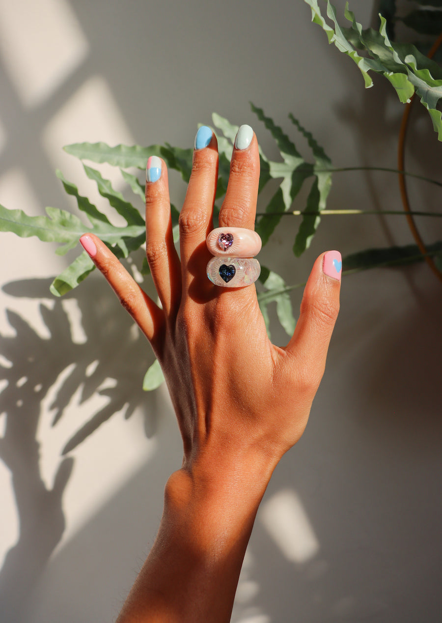 A hand with light brown skin and pastel multicolored nails displays the WALD Berlin SAMPLE Zeena x WALD Blue Heart Ring, a chunky romantic jewelry piece, against green leaves and sunlight.