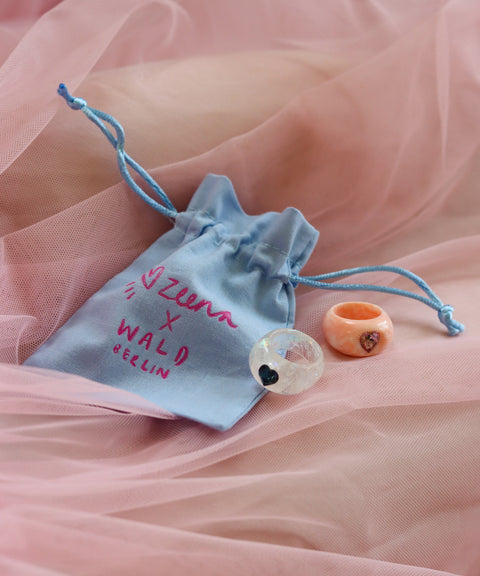 A light blue drawstring pouch labeled "Zeena x WALD Berlin" holds a SAMPLE Zeena x WALD Blue Heart Ring by WALD Berlin, beautifully displayed on pink tulle fabric.