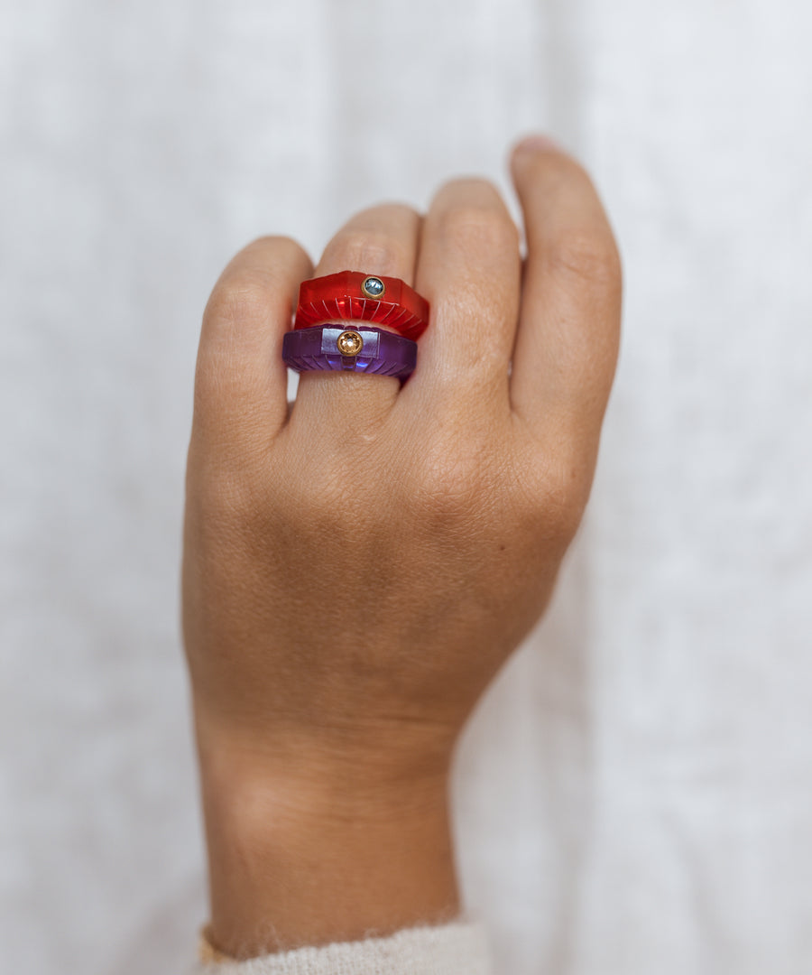 A hand wears two chunky WALD Berlin rings—including the SAMPLE SALE Shining Star Purple Ring—each set with a hand-polished gemstone or Swarovski stone, against a white polyester fabric background made from natural oils.