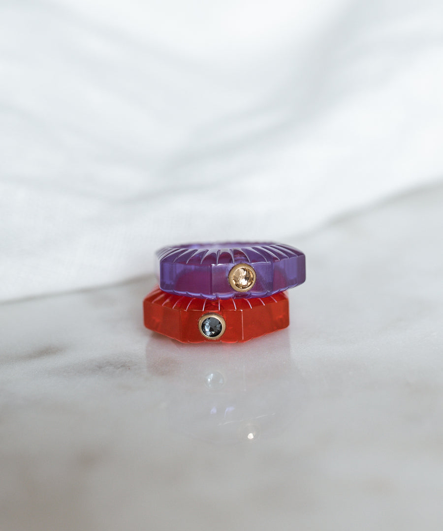 The WALD Berlin SAMPLE SALE Shining Star Purple Ring with a yellow Swarovski stone is stacked with a red ring on marble, set against a white fabric background.